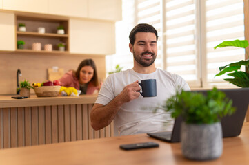 Man working on laptop and drinking coffee, woman cooking