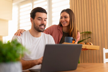 Loving wife bringing breakfast to husband working on laptop