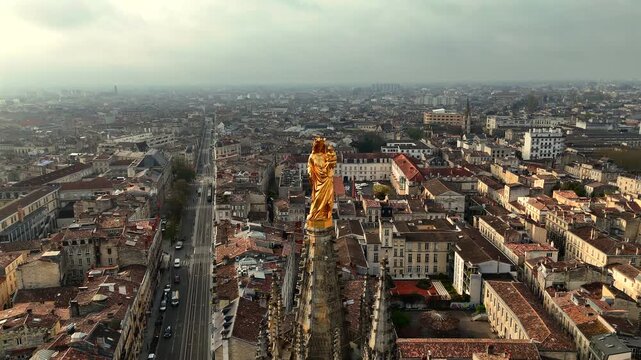 Drone shot of golden Virgin Mary statue atop Pey Berland Tower overlooking straight avenue and historic Bordeaux rooftops during moody weather