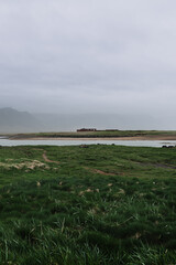 Minimalistic landscape of a small red house near a river in Iceland, surrounded by green fields and misty mountains under a cloudy sky. A calm and cinematic view of Nordic wilderness