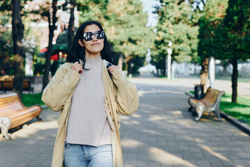 Obraz premium Woman strolling in a sunlit park wearing a beige coat and knit sweater, sunglasses on, casual chic styling in an urban street scene, lifestyle oriented outdoor photography that captures confident,