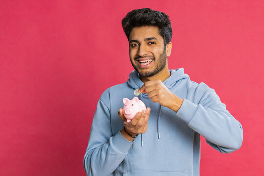 Happy Indian young man holding piggy bank, saving money by dropping coins, planning future expenses and financial security. Smiling Arabian guy investing in savings, isolated on red background - Powered by Adobe