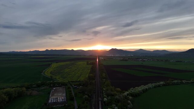 Drone aerial view of railway leading toward mountains at sunset, with a train moving into the distance and sunlight reflecting on rails in a cinematic landscape scene.
