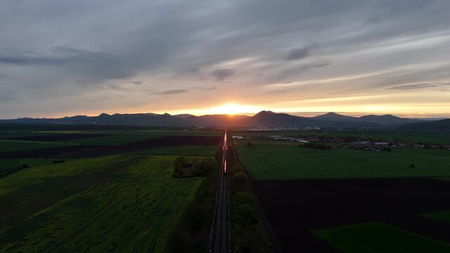Drone aerial view of railway leading toward mountains at sunset, with a train moving into the distance and sunlight reflecting on rails in a cinematic landscape scene.