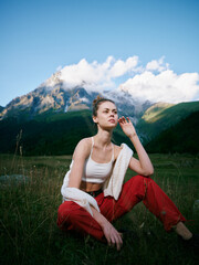 Fototapeta premium Woman sitting on a grassy field with a towel draped over her shoulder, casual white top and red pants, framed by a mountain landscape under a bright blue sky.
