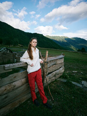 Fototapeta premium Young girl leaning on a rustic wooden fence in a mountainous countryside, wearing a white sweater and red pants, exploring open pasture beneath a bright blue sky.