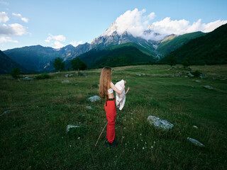 Fototapeta premium Mountains, meadow, hiker, outdoors, expansive landscape with rolling field and distant peaks, a figure in red pants holding a jacket, capturing a peaceful moment in nature