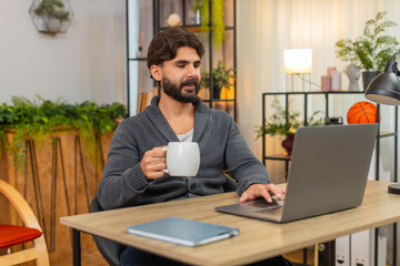 Obraz premium Indian man at home drinks coffee or tea checks tasks on laptop marks done gives thumbs up smiles. Arabian guy at desk reviews list approves progress posts note with joy and feels proud calm mood