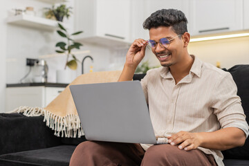 Indian man at home struggles to read tiny text on laptop then puts on glasses and sees clear view. Arabian guy on sofa smiles adjusts frame reads screen lines without strain and continues work task.