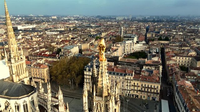 Drone shot of golden Virgin Mary statue atop Pey Berland Tower overlooking straight avenue, tram tracks and historic city rooftops