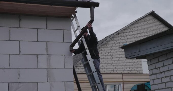 A Caucasian man attaches a gutter. A lone man climbs a ladder to attach a downspout to a house under a gray sky. A man, clutching a toolbox, prepares a downspout from the roof.