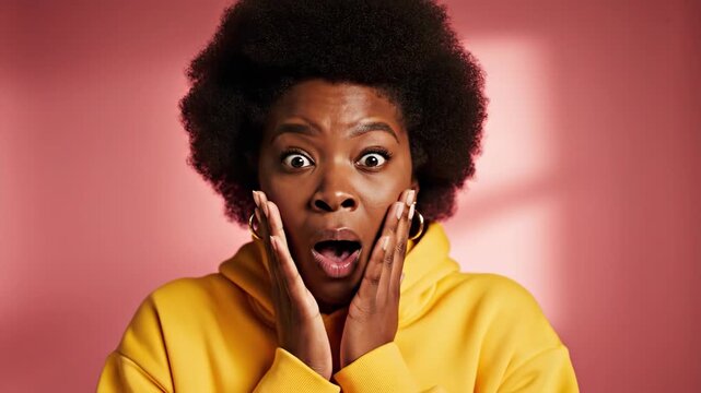 Young African American woman with an afro hairstyle expressing shock and surprise, holding her hands to her face, against a pink background.