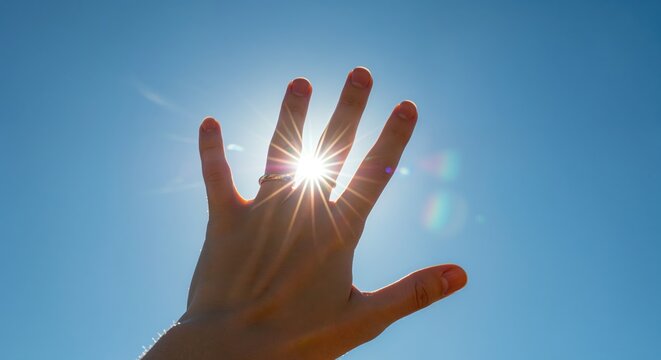 A female hand with a ring reaches up to block the radiant sun starburst, creating a bright lens flare against a vibrant clear blue sky. - Powered by Adobe