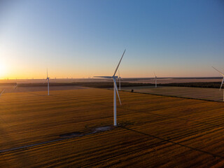 Aerial view of Wind turbines stand across open farmland as the sun sets on the horizon, casting long shadows and warm light over the rural landscape.