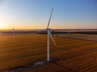 Aerial view of Wind turbines stand across open farmland as the sun sets on the horizon, casting long shadows and warm light over the rural landscape.