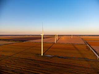 Aerial view of Wind turbines stand across open farmland as the sun sets on the horizon, casting...