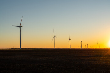 Obraz premium Wind turbines stand across open farmland at sunset, silhouetted against a warm golden sky as the sun drops below the horizon.