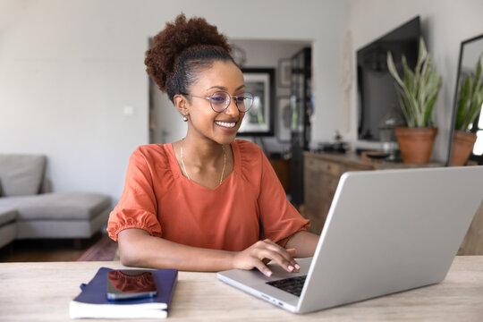 Fototapeta Happy beautiful young African businesswoman in elegant glasses working on online freelance project from home, typing on laptop at workplace table, smiling, using wireless technology