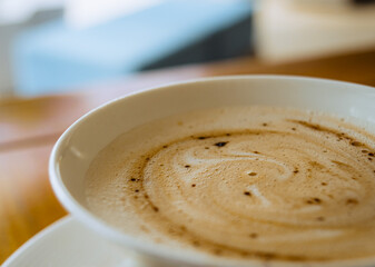 A white ceramic cup filled with dark espresso and textured foam sits on a white saucer atop a grainy wooden tabletop near a blue-painted window.