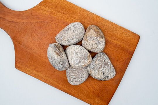 A flat lay of six dried, cracked Pangium edule (kluwek) seeds on a warm-toned wooden cutting board, isolated on a clean white background