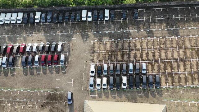 Aerial view taken with a drone of a large showroom parking area displaying neatly arranged new cars on both sides and a walkway in the middle.