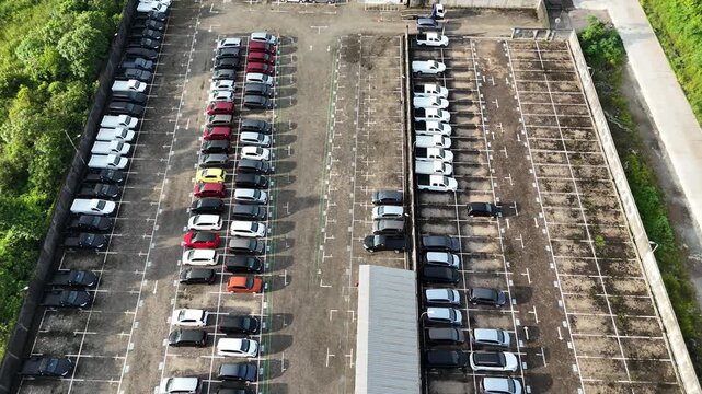 Aerial view taken with a drone of a large showroom parking area displaying neatly arranged new cars on both sides and a walkway in the middle.
