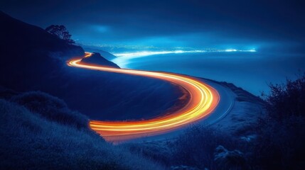 Curved mountain road at night illuminated by bright orange light trails from moving vehicles against a blue misty landscape and distant city lights