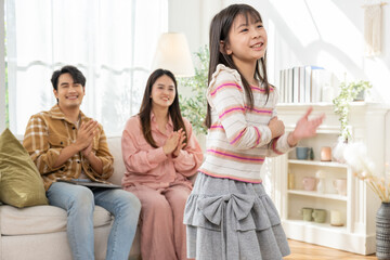 Asian smiling young girl performs dance in bright living room while parents sitting on sofa clap proudly, warm family moment showing joy, parenting, celebration, casual home life