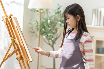 Asian Young girl in apron painting on easel in sunlit living room with plant and lamp, focused and creative expression