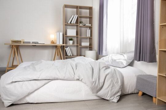 Crisp minimalist bedroom with unmade white bed, gray duvet and pillows by window with soft curtains, wooden desk and open shelving creating calm natural light atmosphere