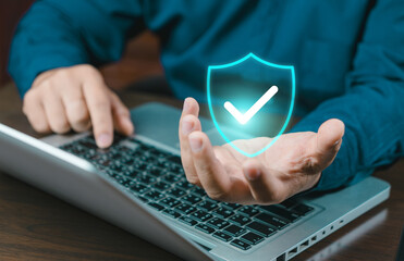 A close-up of a person's hand holding a glowing blue security shield icon with a checkmark above a laptop keyboard, representing data protection and cyber security.