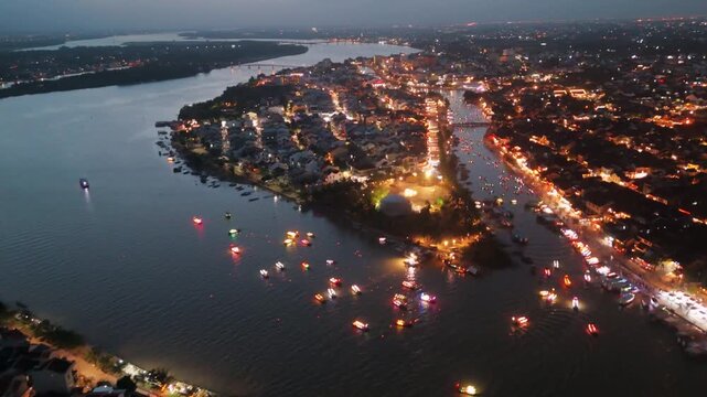 drone fly above Hoi An Ancient Town, Vietnam, at dusk. The Thu Bon River and historic houses are illuminated by glowing traditional lanterns and city lights