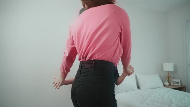 Happy attractive mother dancing to music together with asian daughter at bedroom. Caucasian mom dance or exercise while cute child move to music while jump to bed. Family recreation concept. Pedagogy.