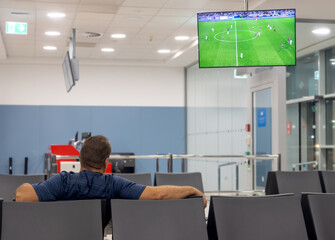 A man sits back in a quiet waiting area at airport terminal, watching a soccer match on a TV