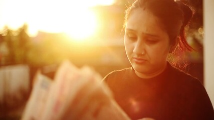 A young adult Indian woman reads a newspaper outdoors during warm golden hour light. The scene reflects daily news reading, awareness, calm moments, and modern lifestyle in India.
