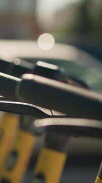 closeup scooter handlebars in sunlight, rental fleet lined on city curb during golden hour, shallow depth of field bokeh highlights brake lever and textured grip, commuter unlocking scooter, delivery