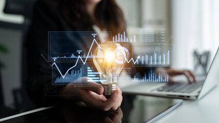 A business professional demonstrates innovation by holding a light bulb while analyzing data trends on a laptop, symbolizing creativity and growth strategy in a modern workspace. Scalp © InfiniteFlow