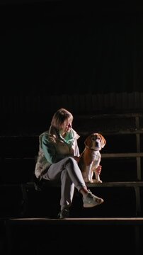 white woman seated on bleachers under single spotlight, beagle companion sitting close, casual jacket and sneakers, nighttime stadium empty rows, contemplative gaze, warm bond highlighted by cinematic