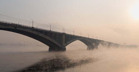 Fototapeta premium An old arched bridge over the Yenisei River, which does not freeze in winter, against the background of foggy sunset in winter in Krasnoyarsk, Russia. Object cultural heritage and a symbol of the city
