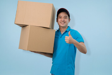 Happy young Asian male courier smiling confidently while carrying two large cardboard delivery boxes and giving a thumbs up, symbolizing approval, excellent service, and reliability