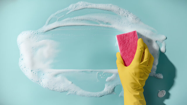 Hand in yellow rubber glove wiping soap foam with sponge on blue background, cleaning concept with copy space