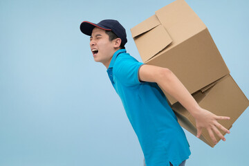 Struggling young Asian man in a blue polo shirt and cap carrying a stack of two large heavy cardboard boxes for packaging grimacing from the effort and weight against a light blue background