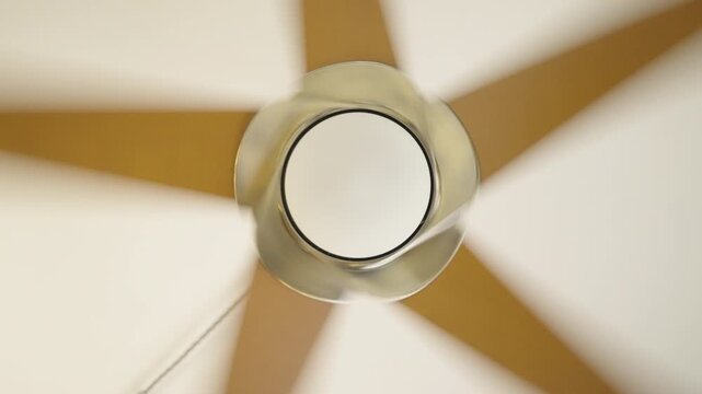 Top-down view of a modern ceiling fan in motion with a metallic hub, five blurred wooden blades, soft lighting, and a dangling pull chain creating a dynamic radial pattern indoors.