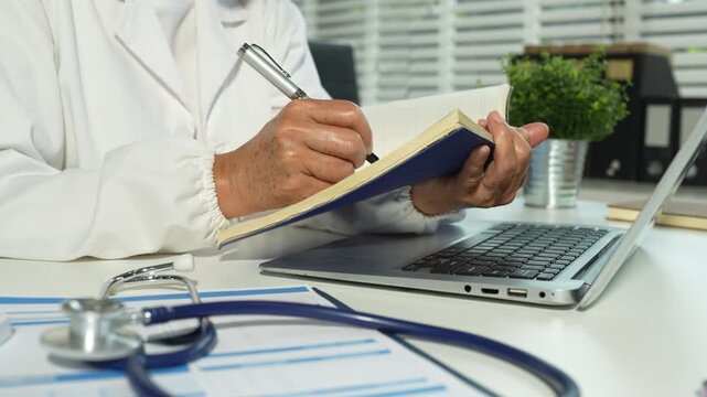 Male medicine doctor, physician or practitioner in lab room writing on blank notebook and work on laptop computer with medical stethoscope on the desk.
