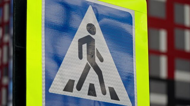 Close-up of a high-visibility pedestrian crossing sign with reflective fluorescent border, highlighting road safety near a zebra crosswalk against blurred urban building background