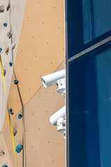 Outdoor climbing wall with safety holds, bright sunlight, and security cameras nearby on a building facade reflecting the sky.