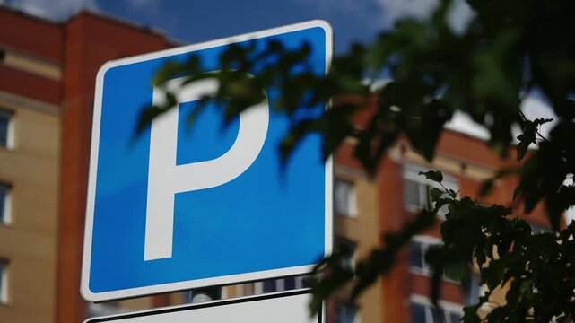 Close-up of a blue parking sign with a bold white P near residential apartment buildings, with leafy branches under a blue sky, showcasing urban street signage and wayfinding