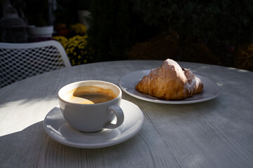 Americano coffee with croissant served at outdoor cafe table with sun light, casual breakfast moment