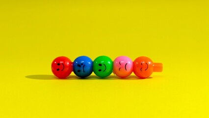 A vibrant hair accessory featuring five colorful beads with different emoticon faces: winking, surprised, smiling, sad, and loving, set against a bright yellow background.