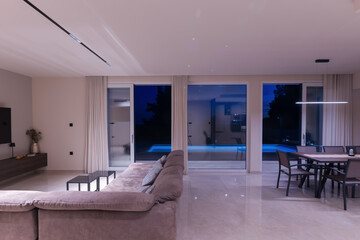  Modern, bright living room featuring a comfortable sofa and panoramic glass doors opening to a stunning private swimming pool and lush garden. Perfect for luxurious indoor-outdoor living